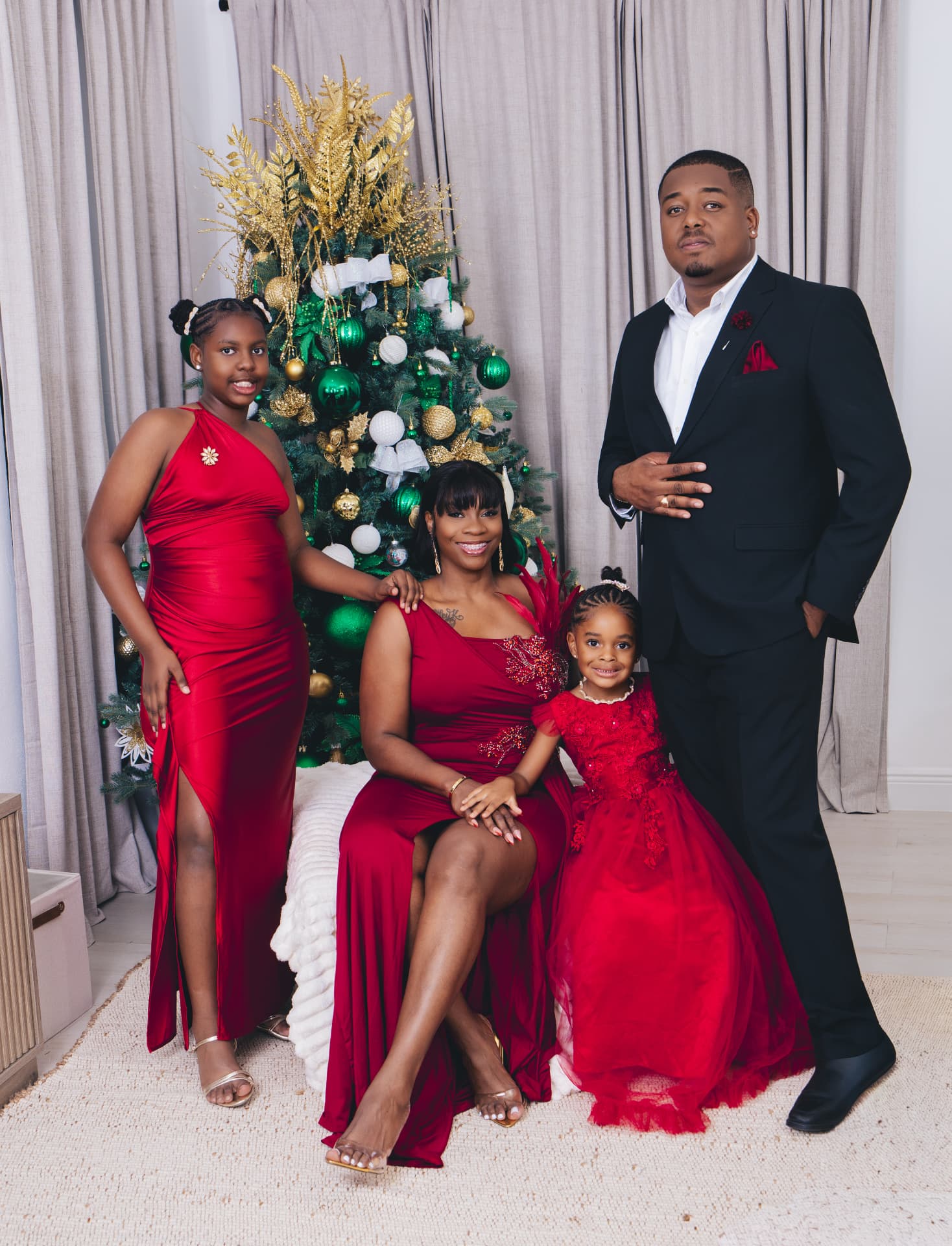 Family of four in red gowns and a black suit posed beside a decorated Christmas tree
