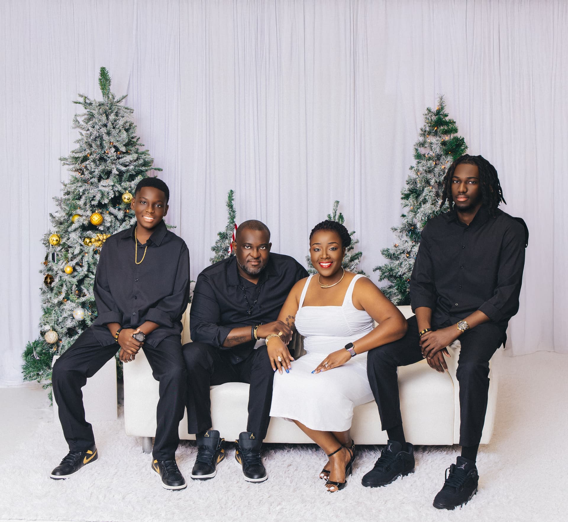 Family of four in black outfits and a white dress seated on a white sofa against white curtains and frosted Christmas trees