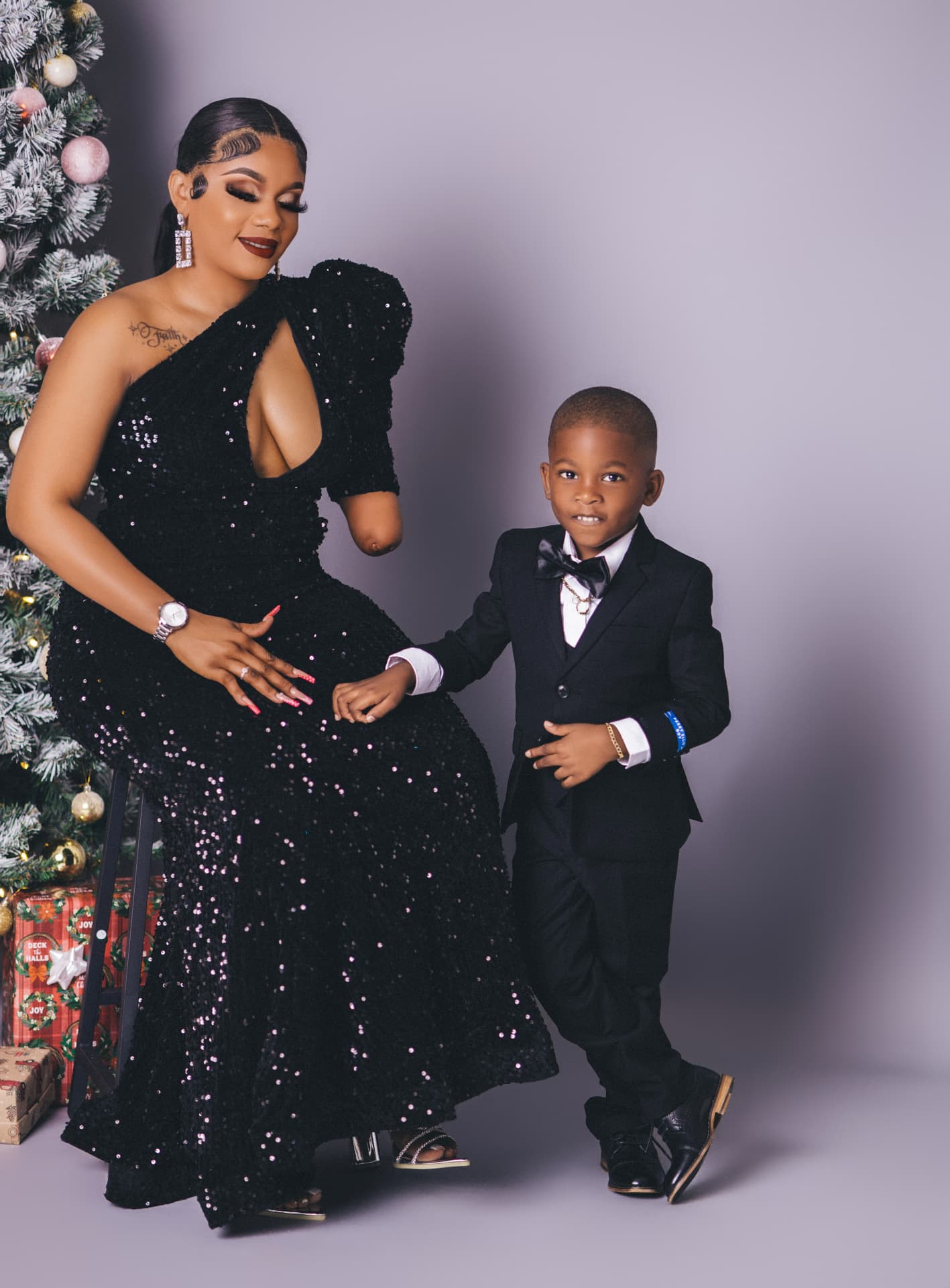 Mother in a sequined gown seated beside her young son in a black suit, next to a frosted Christmas tree with wrapped gifts