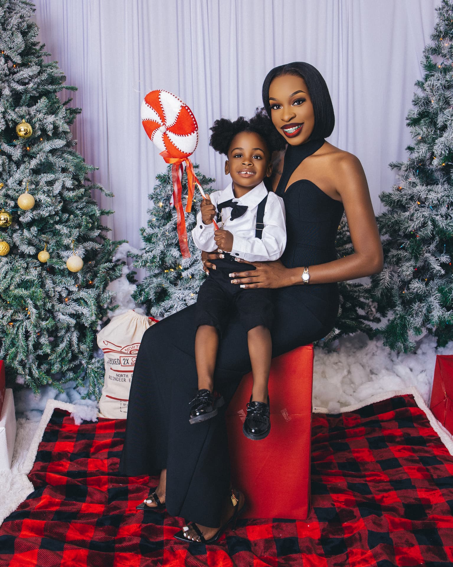 Parent in a black halter dress beside a child in a white shirt, black bowtie and suspenders in a winter studio with white drapes, frosted trees and faux snow