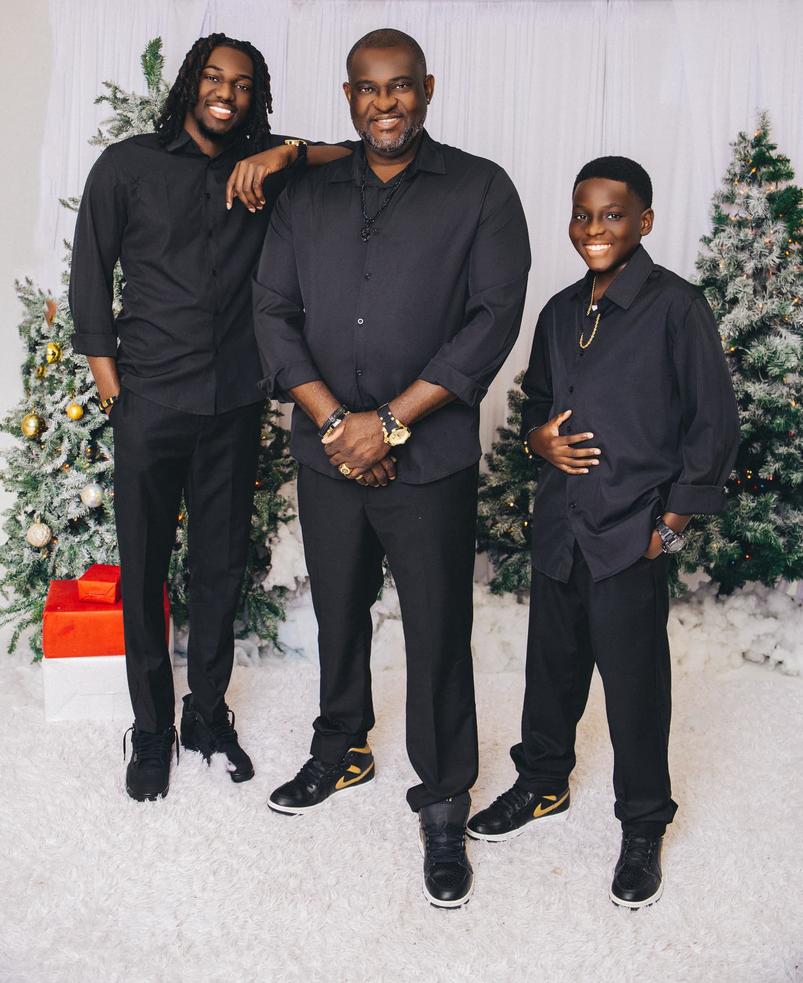 Family of three in matching black shirts and trousers standing on a white carpet before white curtains and frosted Christmas trees