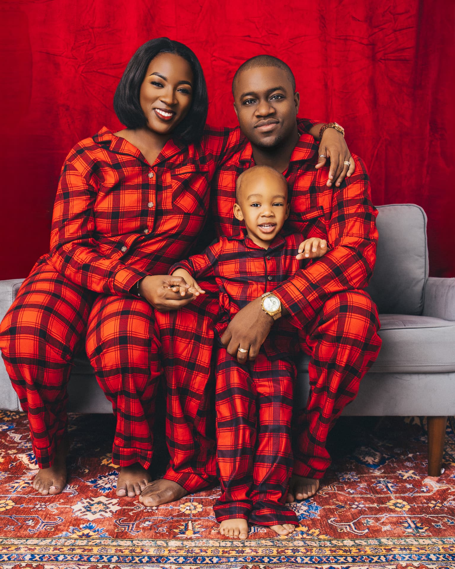 Family of three in matching red and black plaid pajamas seated on a grey couch in front of a red backdrop