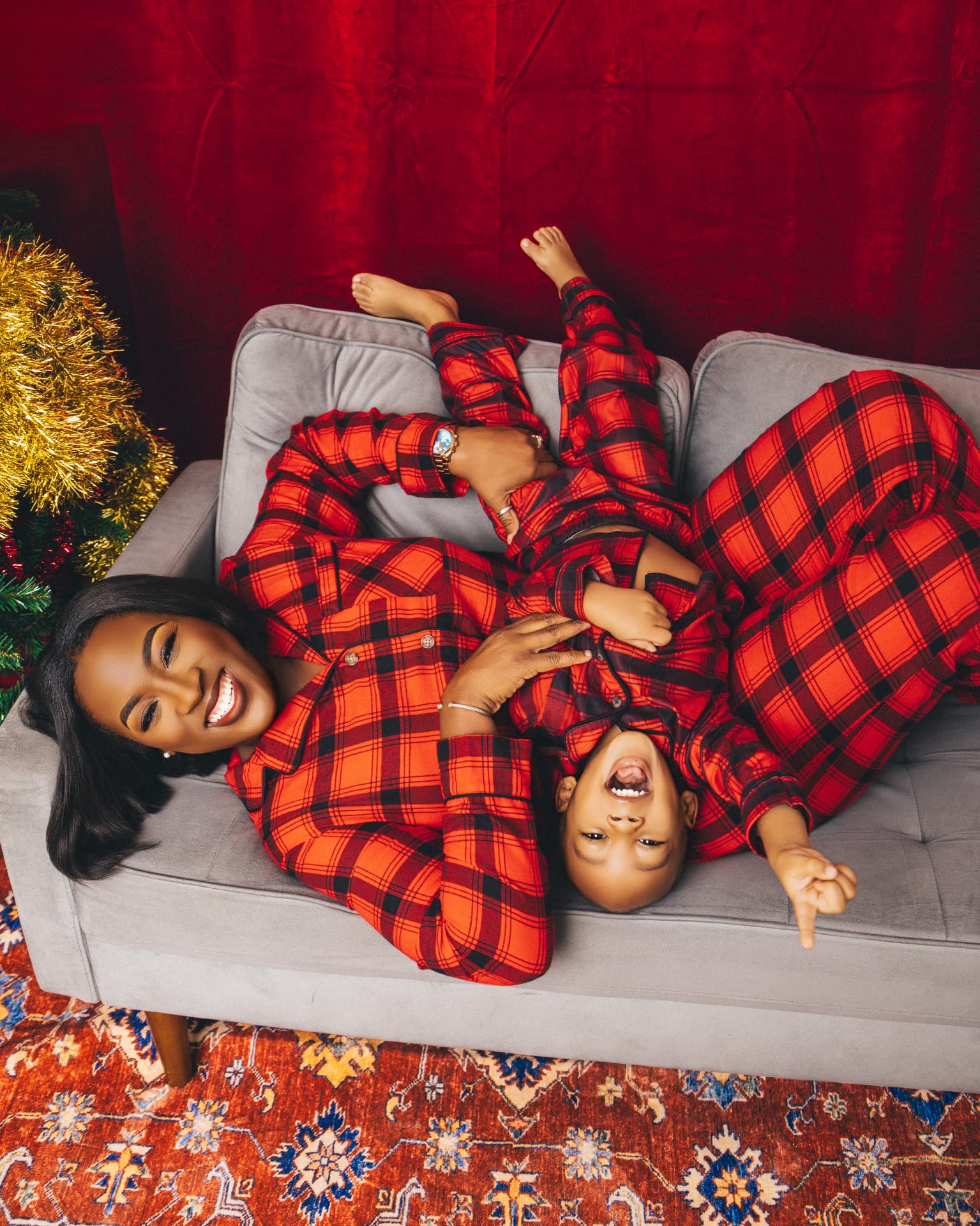 Parent and child in matching red and black plaid pajamas lying together on a grey couch beside a Christmas tree against a deep red backdrop