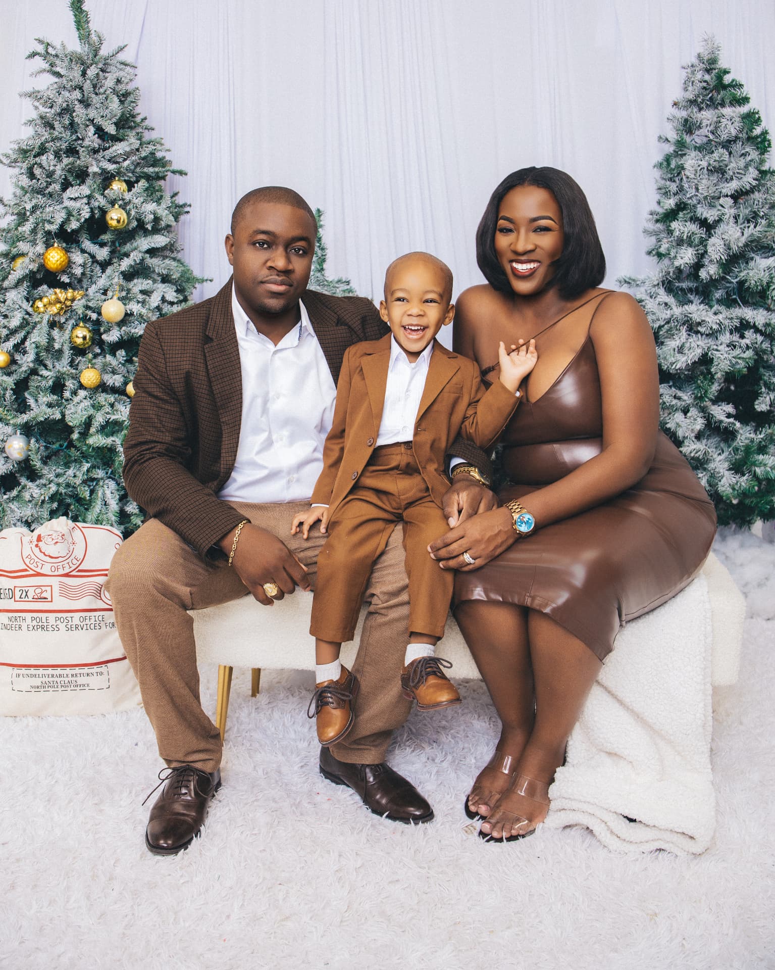 Family of three seated together on a snowy set with frosted pine trees and gold ornaments