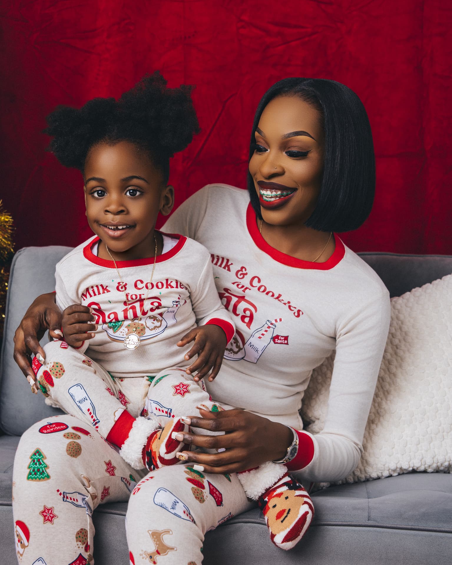 Parent and child in matching white and red Milk and Cookies for Santa pajamas sitting on a grey couch against a red backdrop
