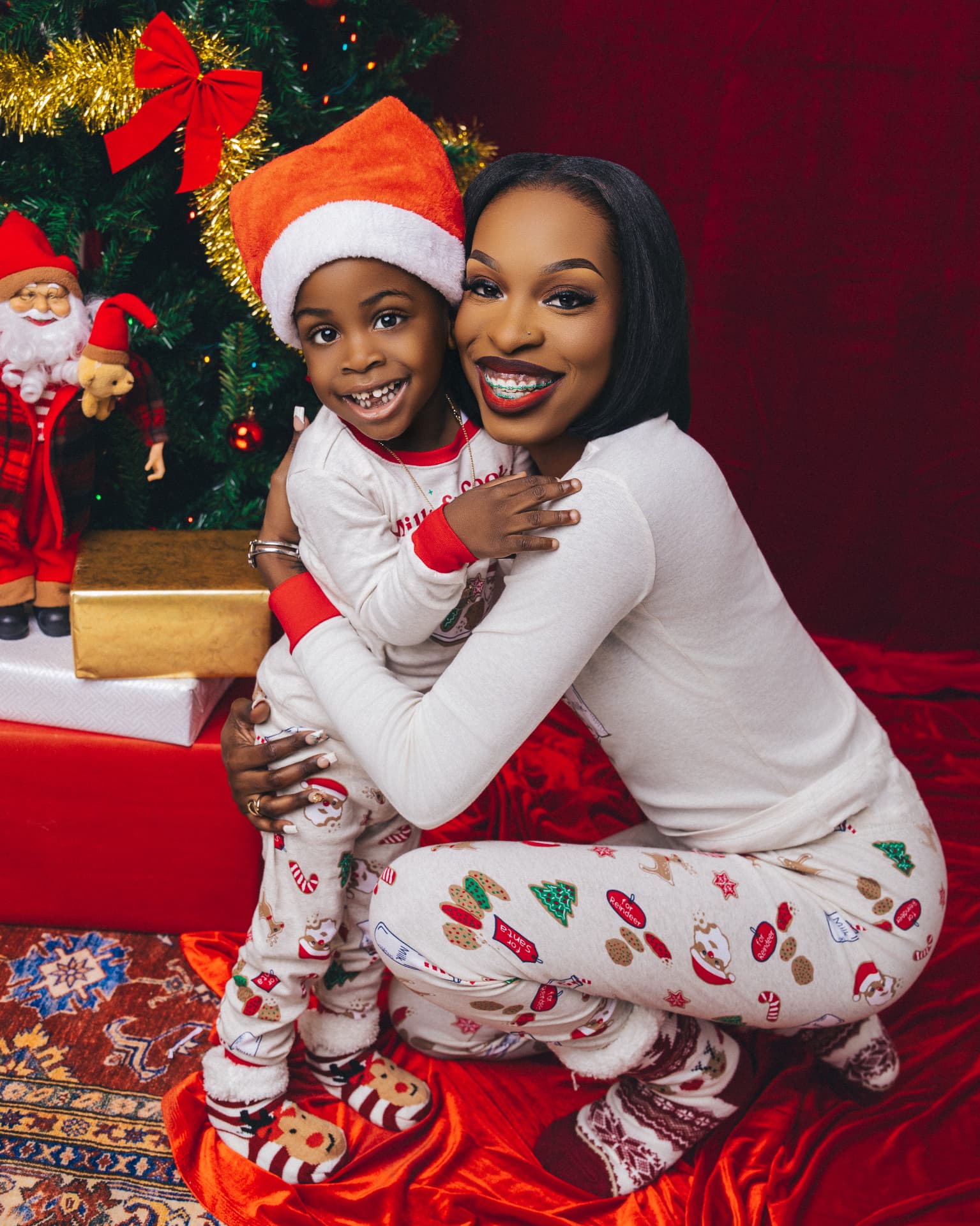 Parent and child in matching white holiday-print pajamas and a Santa hat beside a decorated Christmas tree with gifts on a red backdrop