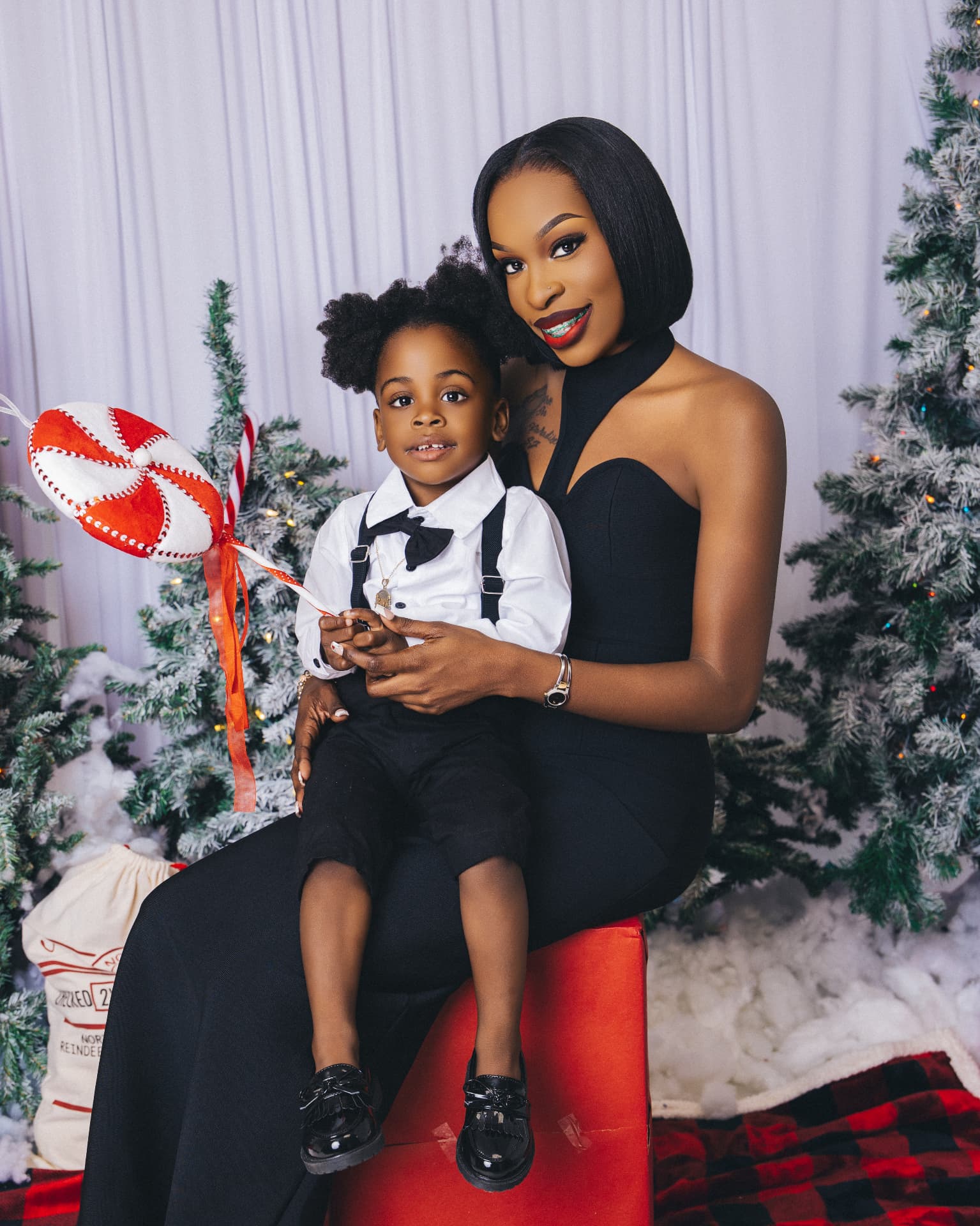 Parent in a black dress and child in a white shirt with bowtie and suspenders posed against a white drape framed by flocked Christmas trees